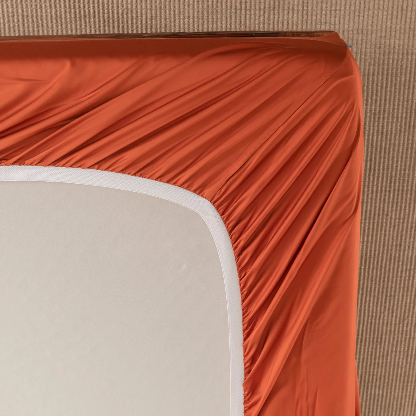 A close-up view of a mattress corner being covered by a fitted sheet in a rich rust color on a beige textured surface. The sheet is partially pulled over the mattress, showing gathered fabric at the edge.