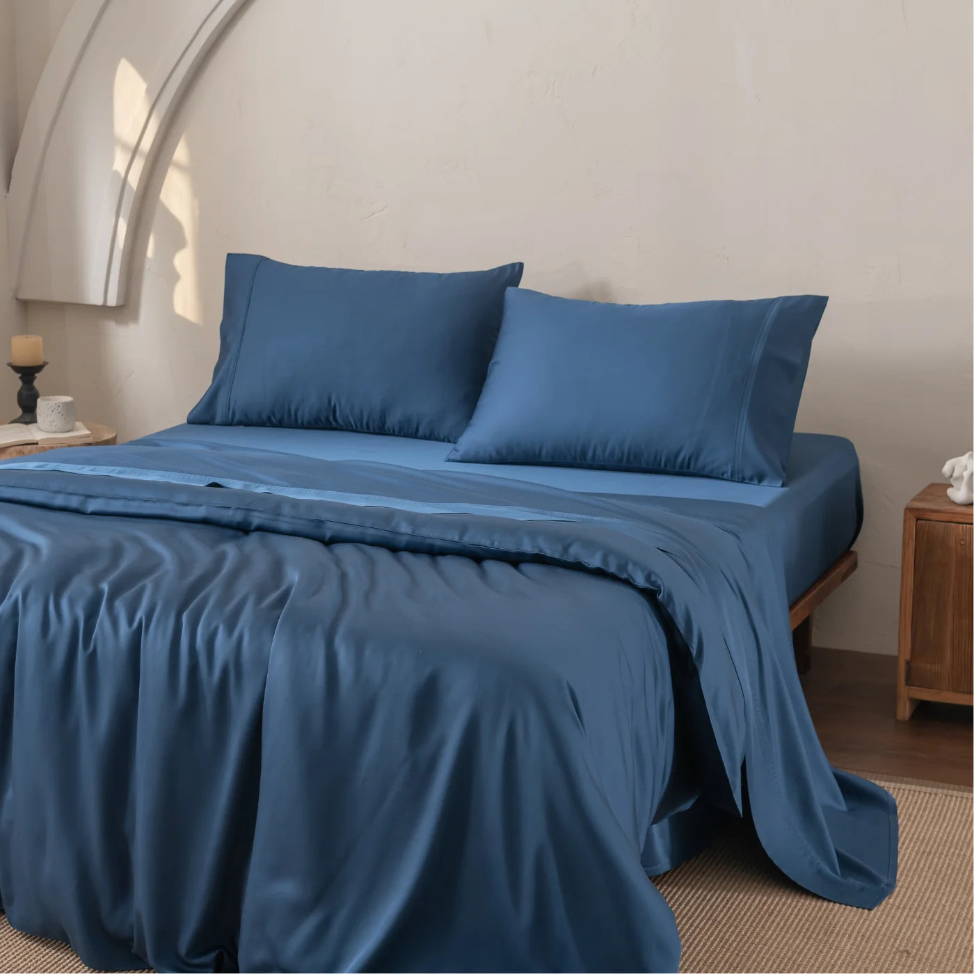 A neatly made bed with deep blue satin sheets and pillowcases, set against a cream-colored wall. A wooden nightstand with a small white vase and a candle is next to the bed. Natural light brightens the room.