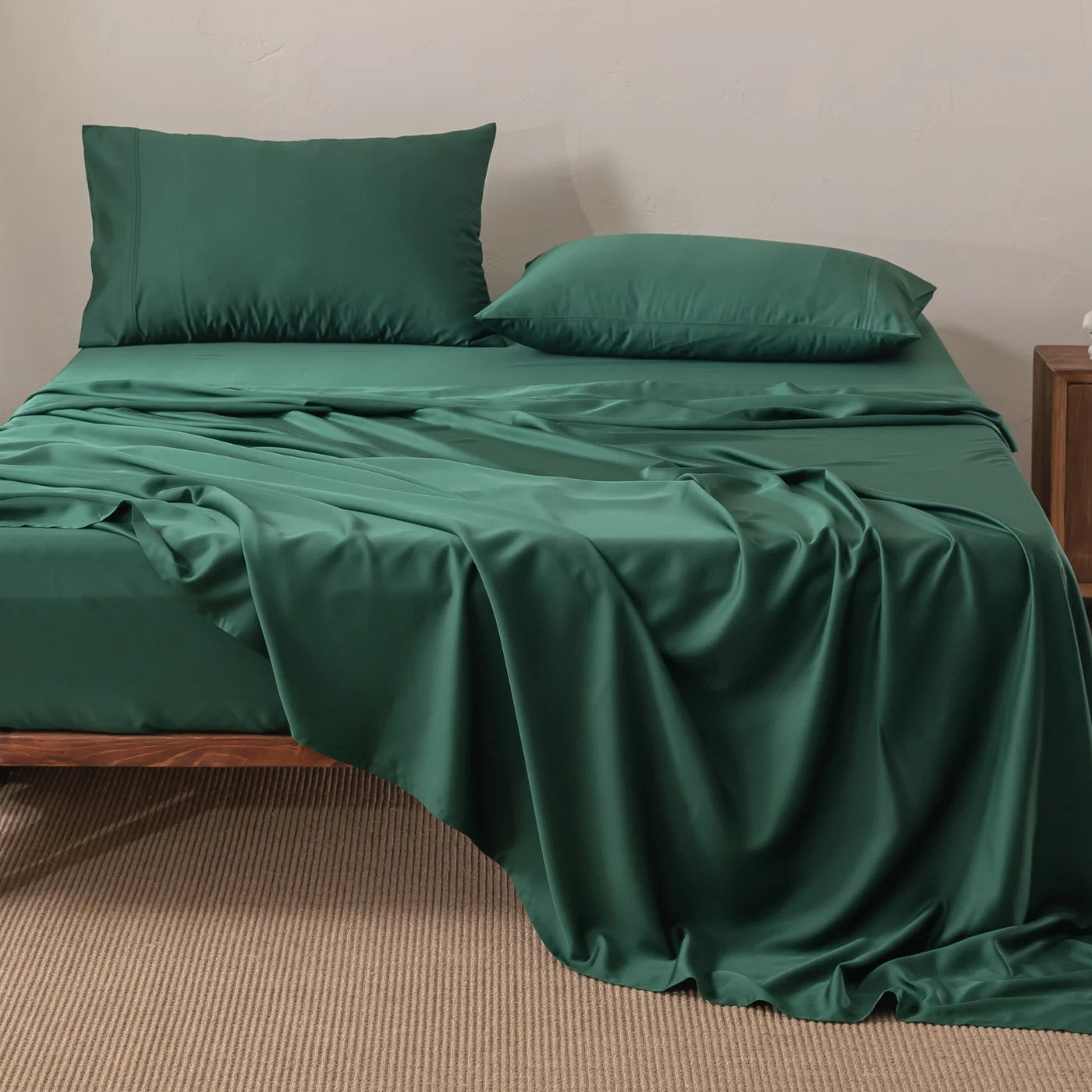 A neatly made bed with a wooden frame, green sheets, and two green pillows. The top sheet is partially draped over the side. A nightstand is visible to the right, and the floor is covered with a beige carpet.