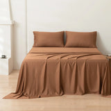 A neatly made bed with matching brown sheets and two pillows, draped so the fabric pools slightly onto a light wooden floor, against a plain white wall in a minimal, modern room.