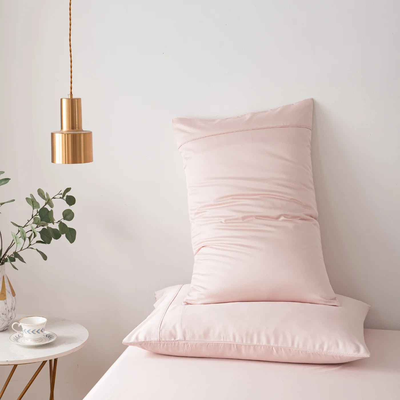 A neatly made bed with light pink bedding, featuring two pillows stacked upright. Nearby, a gold pendant lamp hangs above a round white side table holding a teacup and saucer, a small plate, and a vase with green leaves.