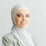 A woman wearing a light gray hijab and a light green blazer smiles confidently while standing in front of a white brick wall.
