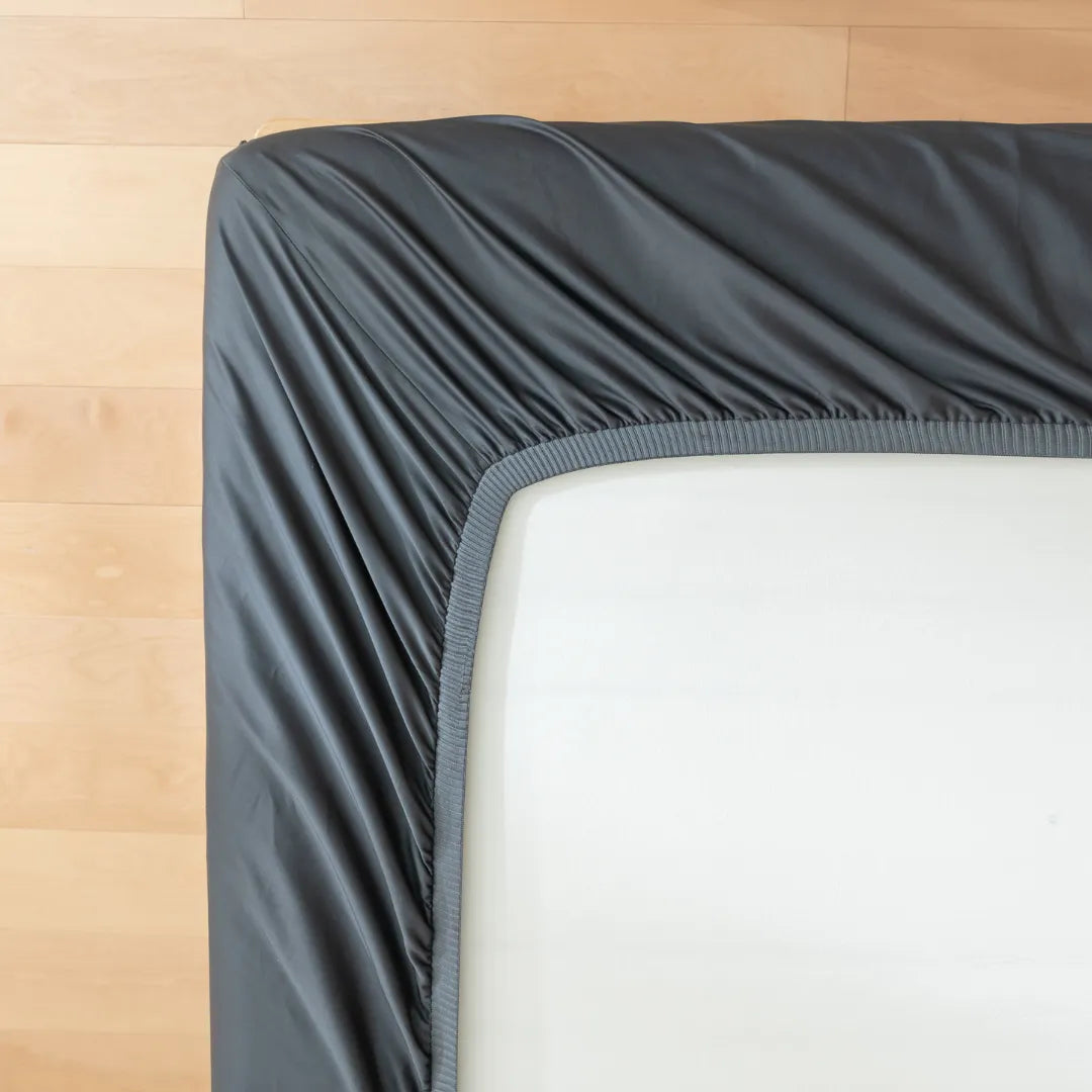 A close-up of a mattress corner with the Linenly Luxe Cotton Sateen Fitted Sheet in charcoal, partially pulled over it on a light wooden floor—ideal for lovers of modern bedding.