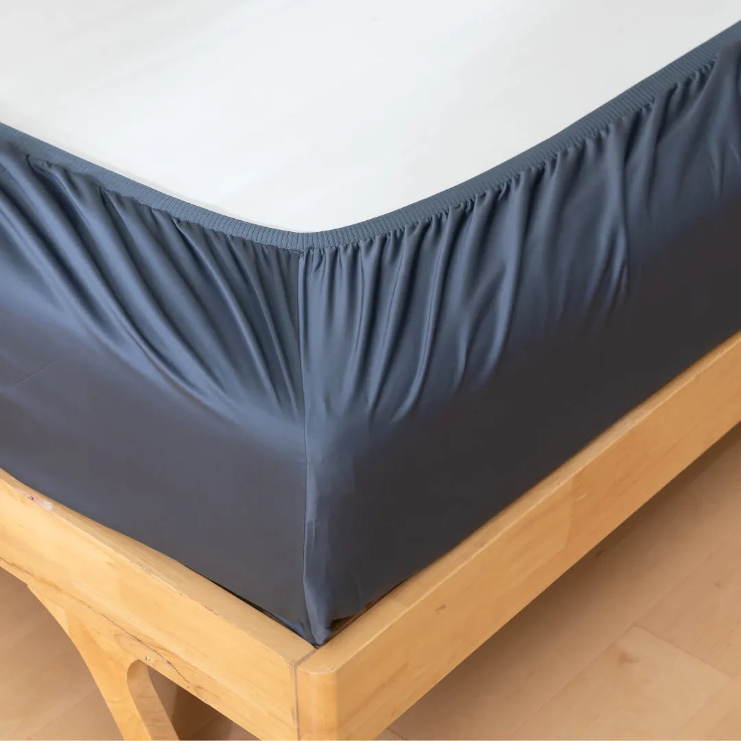 Close-up of the Linenly Luxe Cotton Sateen Fitted Sheet - Midnight, its elastic edges wrapping snugly around the wooden bed frame’s mattress for a smooth, wrinkle-free finish.