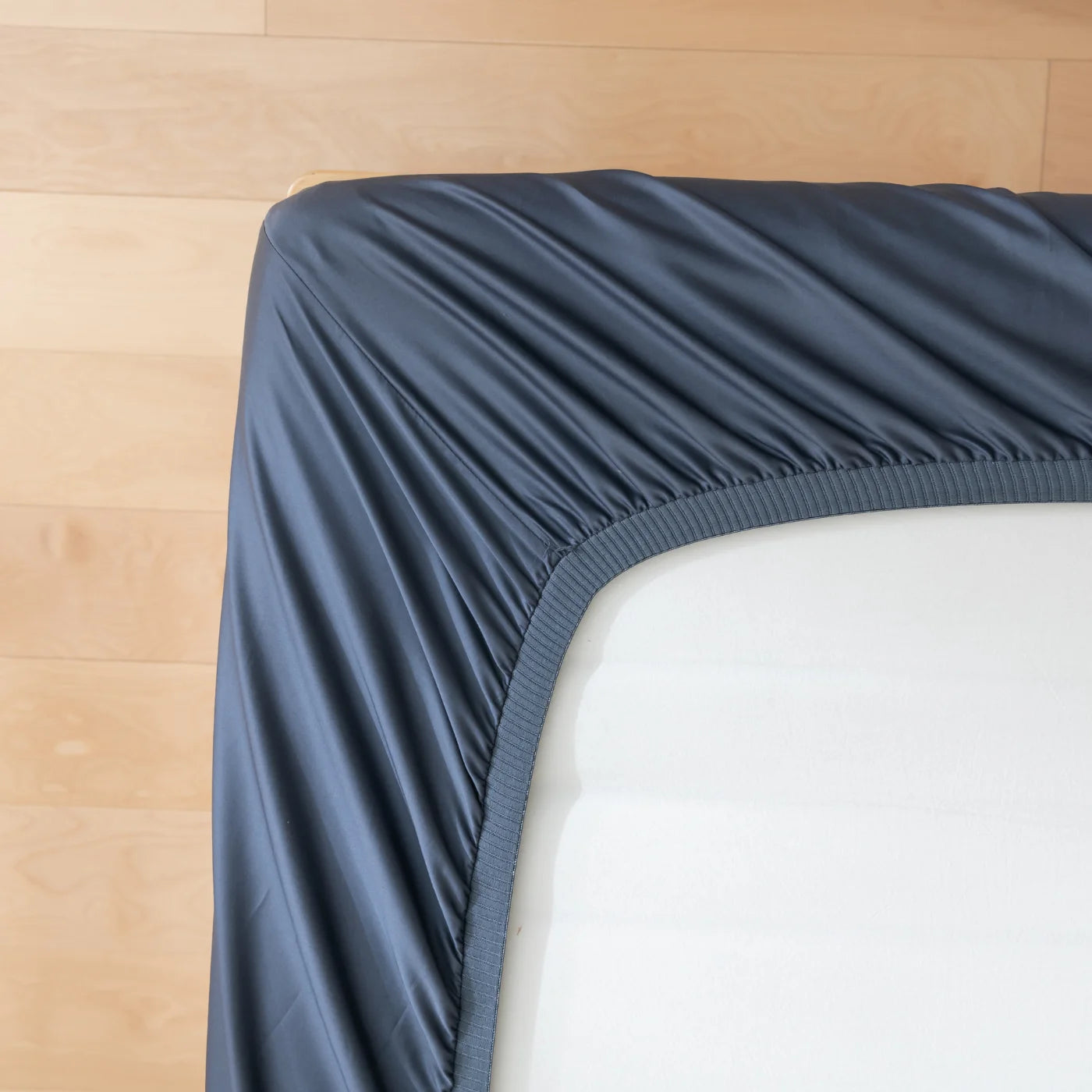 A close-up of a fitted dark gray bedsheet partially pulled over the corner of a white mattress, with a light wooden floor in the background.