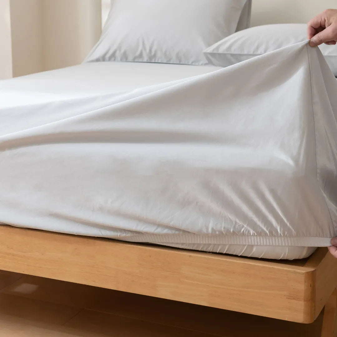 A hand pulls the Linenly Luxe Cotton Sateen Fitted Sheet in Silver over a mattress corner on a light wooden bed, with white pillows and bedding in a sunlit room.