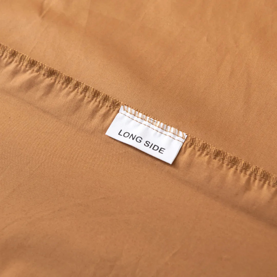 A close-up of Linenly’s Luxe Cotton Sateen Fitted Sheet in terracotta shows a “LONG SIDE” label stitched into the seam, highlighting this cozy bedding essential.