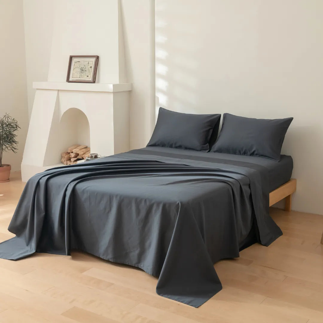 A neatly made bed with Linenly’s Luxe Cotton Sateen Flat Sheet in Charcoal and two pillows sits in a bright, minimal bedroom featuring a fireplace with logs, framed picture, potted plant, and light wooden floors.