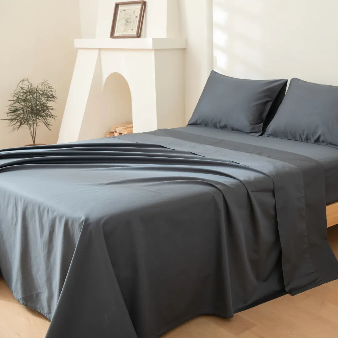 A neatly made bed features the Linenly Luxe Cotton Sateen Flat Sheet in Charcoal, with matching pillowcases, set in a bright room with light walls, a potted plant, and a white decorative fireplace in the background.