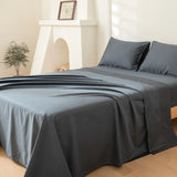 A neatly made bed features the Linenly Luxe Cotton Sateen Flat Sheet in Charcoal, with matching pillowcases, set in a bright room with light walls, a potted plant, and a white decorative fireplace in the background.