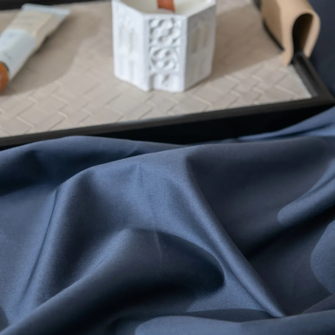 Close-up of the Linenly Luxe Cotton Sateen Flat Sheet in Midnight, its soft dark blue fabric draped in gentle folds, with a white candle and small tube on a beige patterned tray in the background.