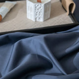 Close-up of the Linenly Luxe Cotton Sateen Flat Sheet in Midnight, its soft dark blue fabric draped in gentle folds, with a white candle and small tube on a beige patterned tray in the background.