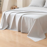 A neatly made bed features the Luxe Cotton Sateen Flat Sheet in Silver by Linenly and white pillows, with part of the sheet elegantly draped on a light wooden floor in a minimal, cozy room. Firewood is stacked on a shelf in the background.