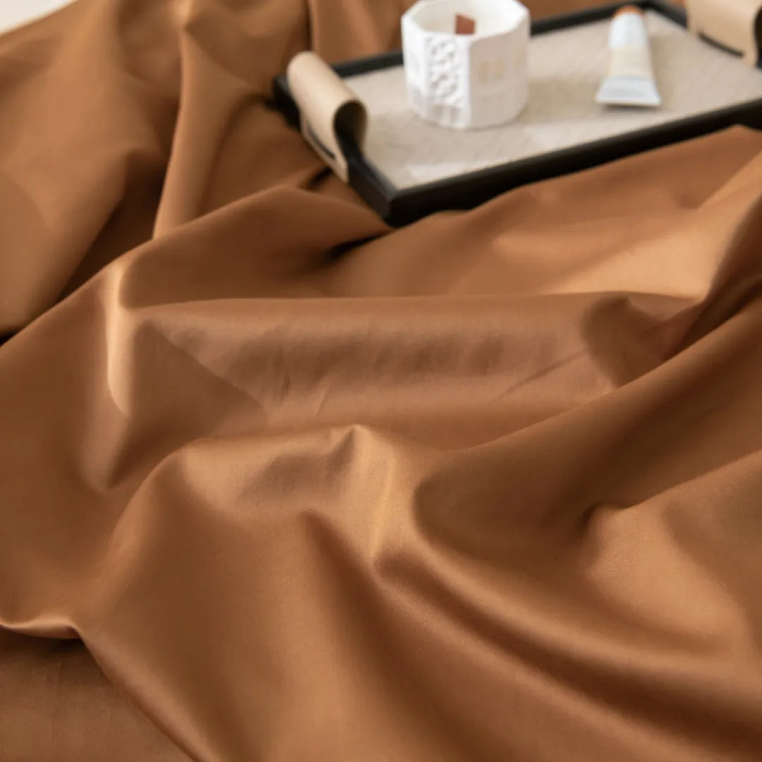 The Luxe Cotton Sateen Flat Sheet in Terracotta by Linenly is smoothly spread with gentle folds, while a tray in the background holds a rolled-up scroll, white geometric candle, and small tube, all slightly out of focus.