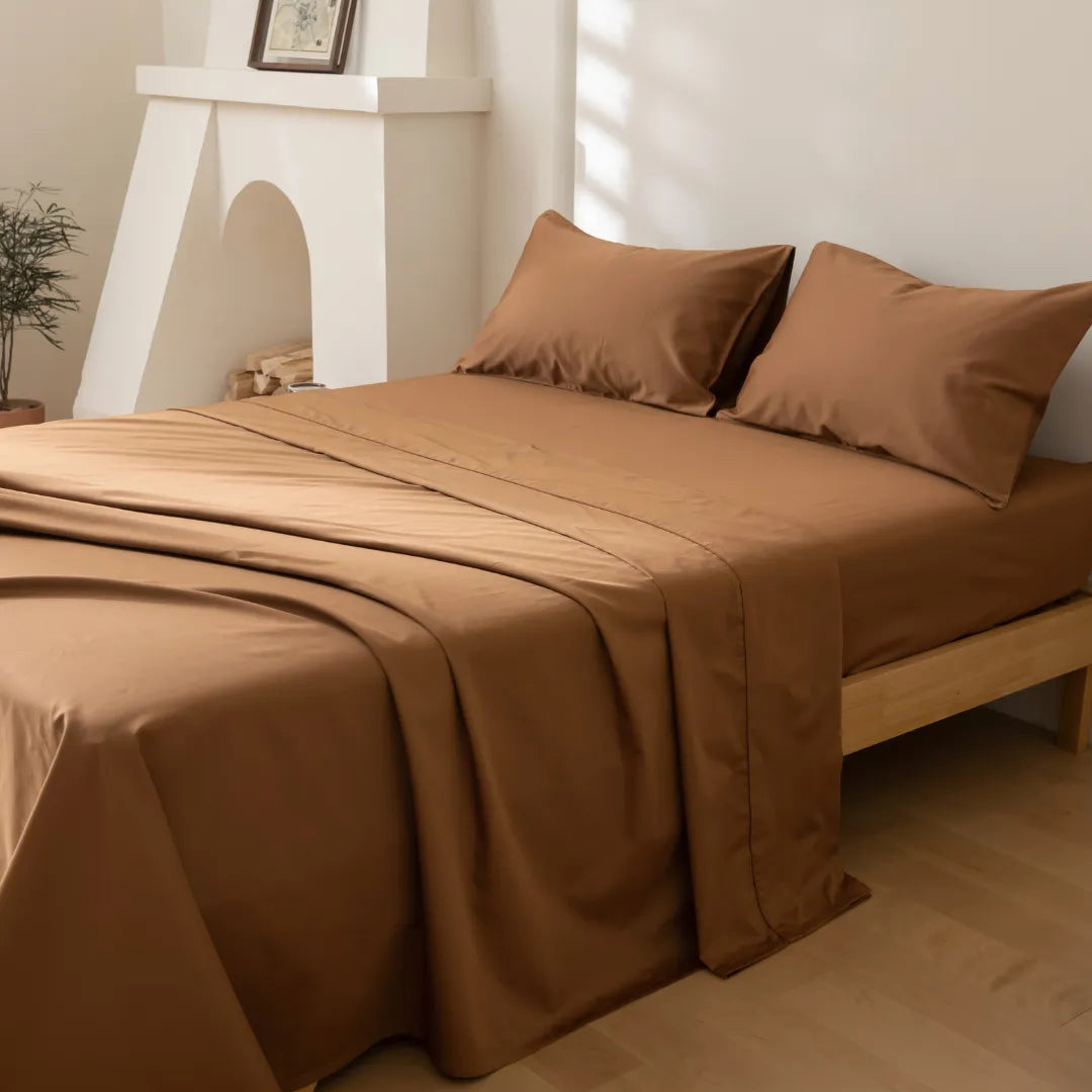A minimalistic bedroom features a bed dressed in Linenly's Luxe Cotton Sateen Flat Sheet in Terracotta with two matching pillows, long staple cotton fabric, light wood floors, white walls, soft natural light, a plant, and framed picture.