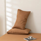 Two brown pillows rest against a light wall; one leans on the other, which lies flat on the Linenly Luxe Cotton Sateen Flat Sheet in Terracotta. A cup, saucer, and magazine are nearby as soft light casts gentle shadows on the wall.