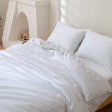 A neatly made bed with the Linenly Luxe Cotton Sateen Flat Sheet in white and matching pillows sits by a stacked firewood fireplace in a bright, minimalistic room.