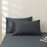 Two Linenly Luxe Cotton Sateen Pillowcases in Charcoal rest against a matching sheet, with sunlight casting soft shadows on the neutral wall. A small tray with a cup and saucer sits at the bed's edge.