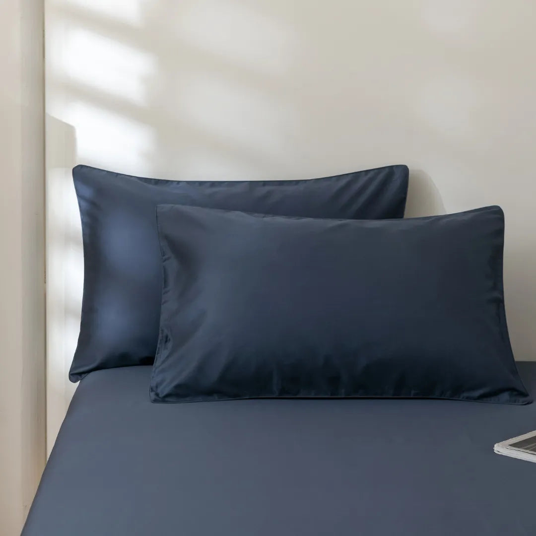 Two Luxe Cotton Sateen Pillowcases in Midnight by Linenly are arranged side by side on a matching sheet, set against a light wall with gentle natural light, highlighting their rich dark blue hue.