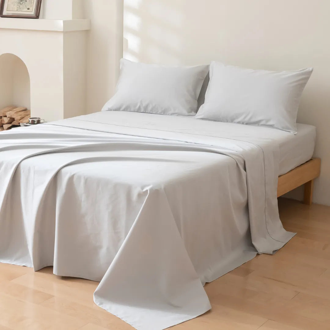 A neatly made bed features Linenly's Luxe Cotton Sateen Pillowcase Set in Silver, with light gray sheets draped over a wooden frame and resting on a light wood floor against a white wall.