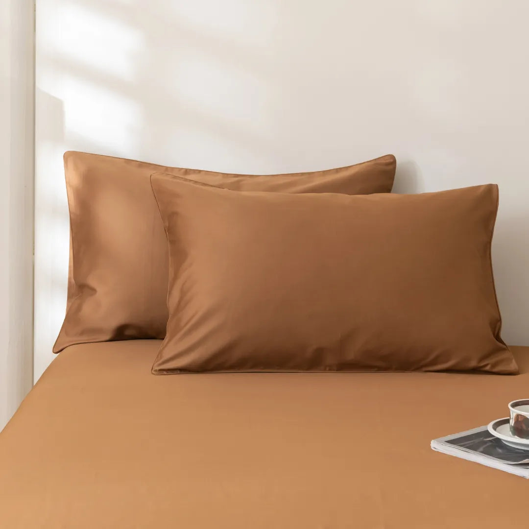 Two Luxe Cotton Sateen Pillowcases in Terracotta by Linenly are set against a matching bedsheet on a neatly made bed. Sunlight casts soft shadows behind, with a book and cup placed on the lower right bedside corner.