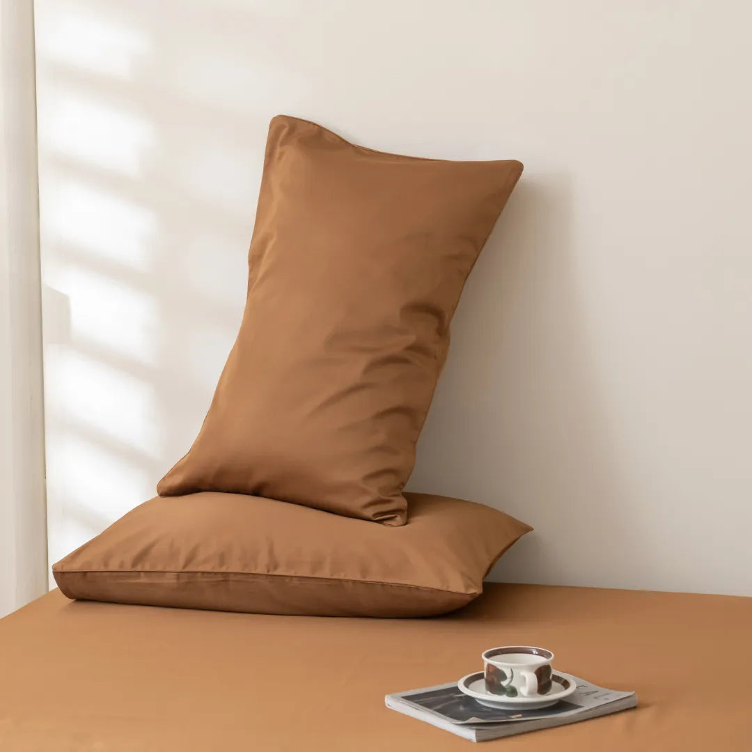 Two Linenly Luxe Cotton Sateen Pillowcase Sets in terracotta are stacked against a light wall on a matching brown surface. In front, a cup and saucer sit on a magazine while soft natural light casts gentle shadows.