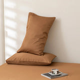 Two Linenly Luxe Cotton Sateen Pillowcase Sets in terracotta are stacked against a light wall on a matching brown surface. In front, a cup and saucer sit on a magazine while soft natural light casts gentle shadows.