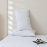 Two Linenly Luxe Cotton Sateen Pillowcases in white are stacked on a bed against a light wall. Sunlight creates soft shadows, while a cup of coffee and magazine sit atop the bed in the foreground.