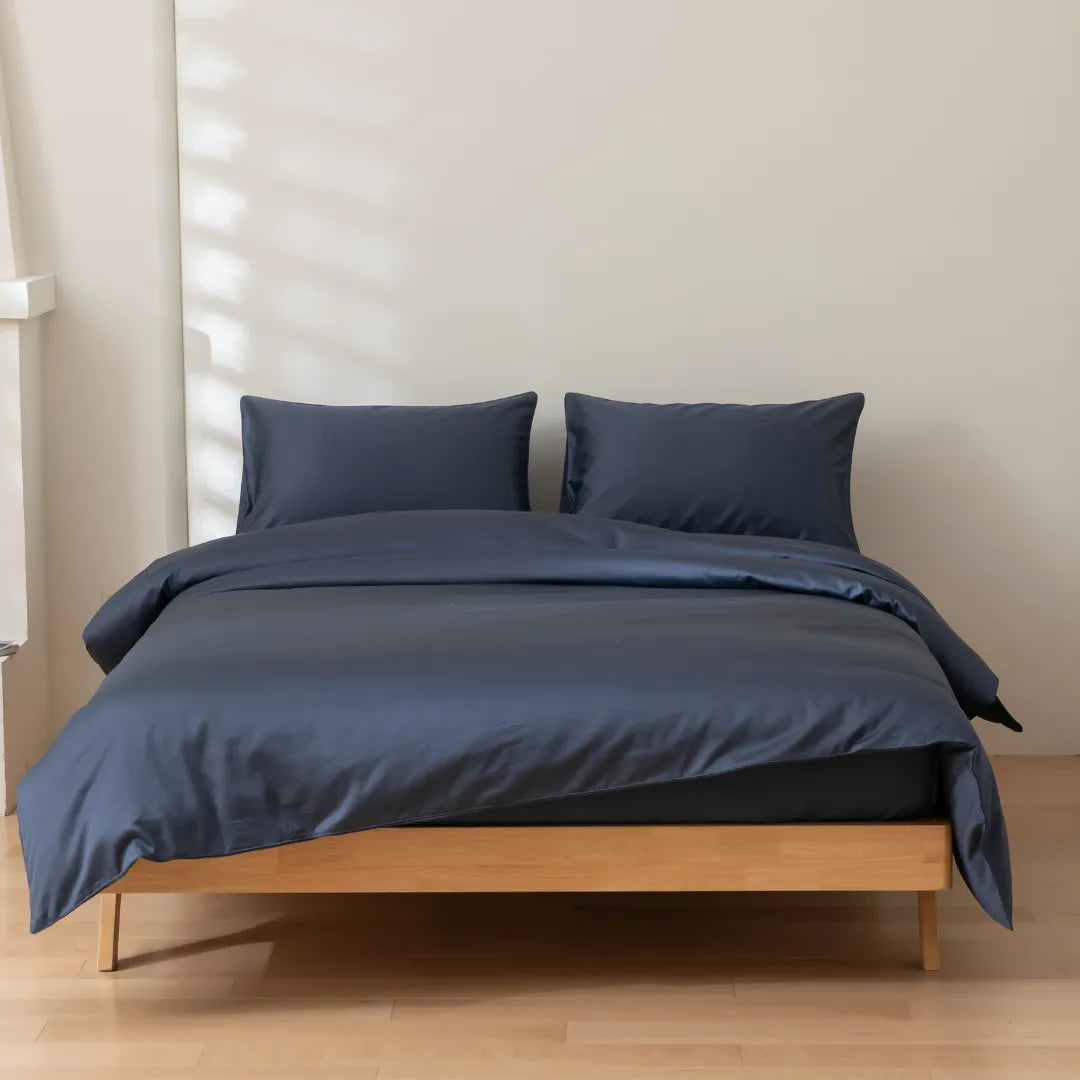 A neatly made bed features Linenly's Luxe Cotton Sateen Quilt Cover in Midnight and two matching pillows, resting on a light wooden frame in a minimalistic room with pale walls and wooden flooring.