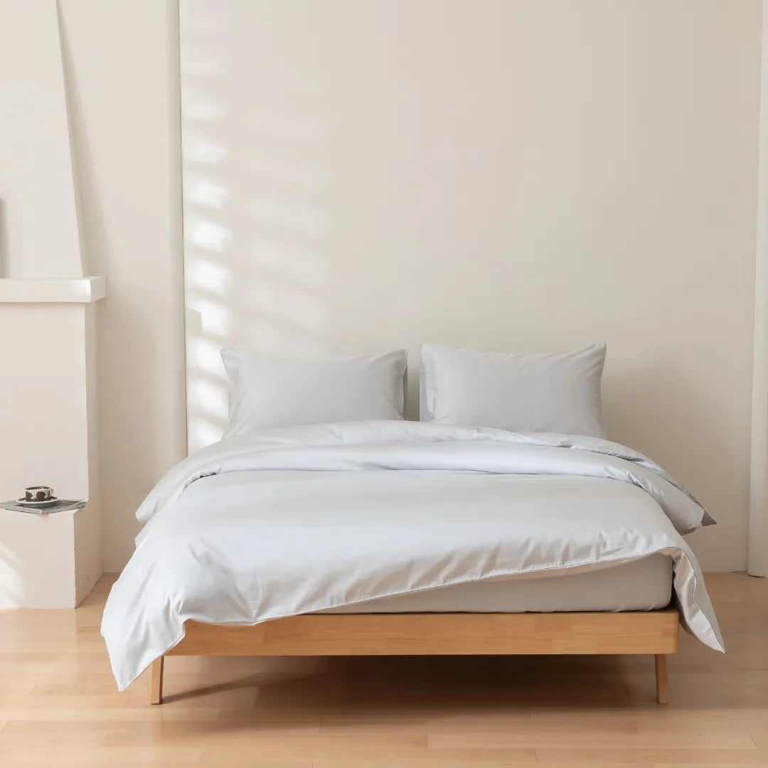 A neatly made bed featuring the Linenly Luxe Cotton Sateen Quilt Cover in Silver and two pillows rests against a plain light wall on a wooden floor. Soft natural light casts gentle shadows, adding to the calm, minimalist ambiance.