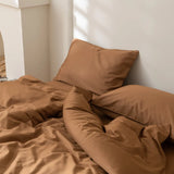 A bed with Linenly's Luxe Cotton Sateen Quilt Cover in terracotta and two pillows is set against a white wall, with sunlight highlighting the warm, inviting look of the tan bedding.