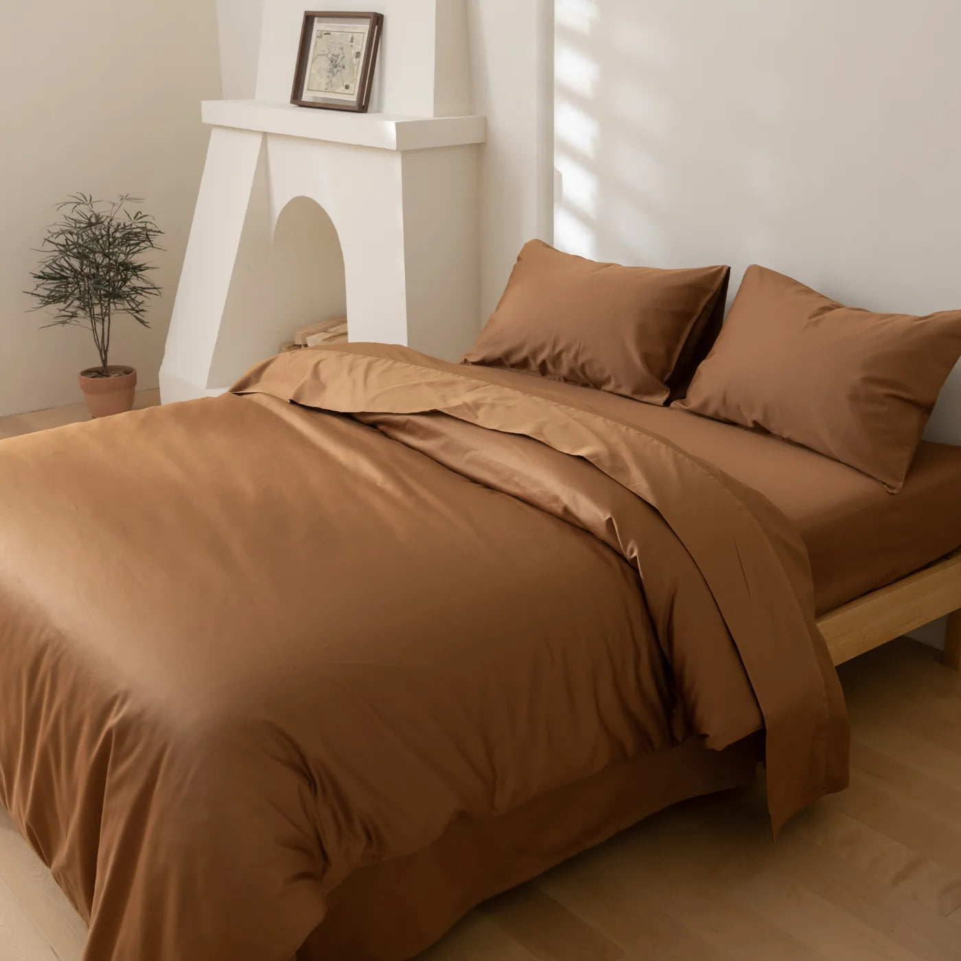A modern bedroom with a neatly made bed covered in brown sheets and pillowcases, a small potted plant, and a white fireplace mantel with a framed picture, all set against light-colored walls and floor.