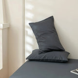 Two dark gray pillows rest atop the Linenly Luxe Cotton Sateen Sheet Set in Charcoal, with soft natural light casting shadows on a light-colored wall in the background.