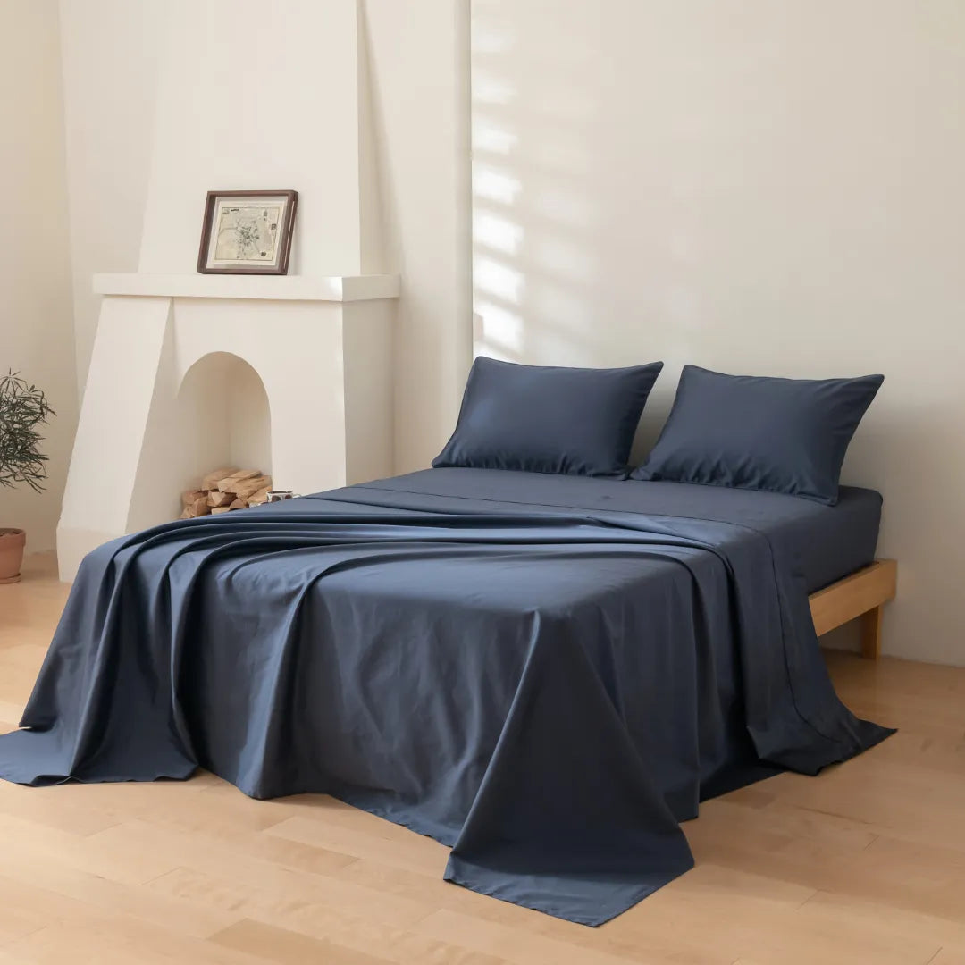 A minimalist bedroom features a neatly made bed dressed in Linenly’s Luxe Cotton Sateen Sheet Set in Midnight, with two matching pillows. Sunlight streams in, highlighting a small mantle with a framed picture and stacked firewood.