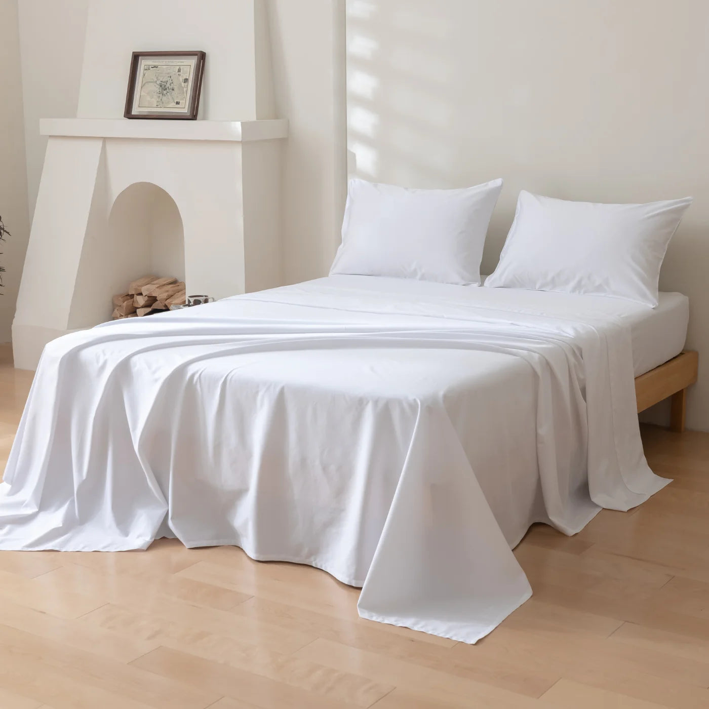 A neatly made bed with white sheets and two pillows is in a bright room with light wooden floors. The bed sits next to a white fireplace holding a framed picture. Sunlight streams in, casting shadows on the wall.