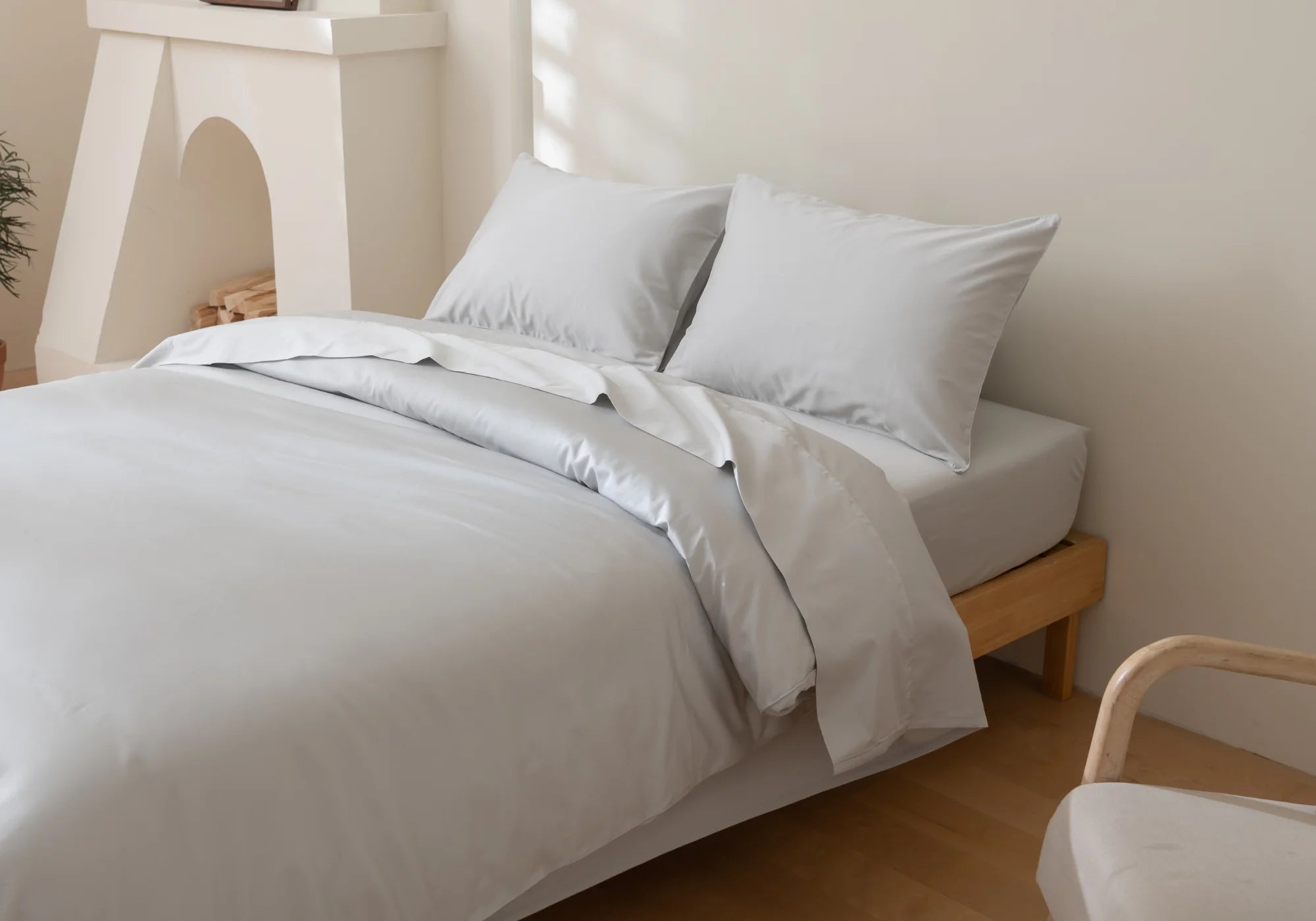 A neatly made bed with light gray sheets, pillowcases, and duvet cover in a minimal, modern bedroom with light walls, wooden bed frame, and a soft chair beside the bed. Natural light streams in from the side.
