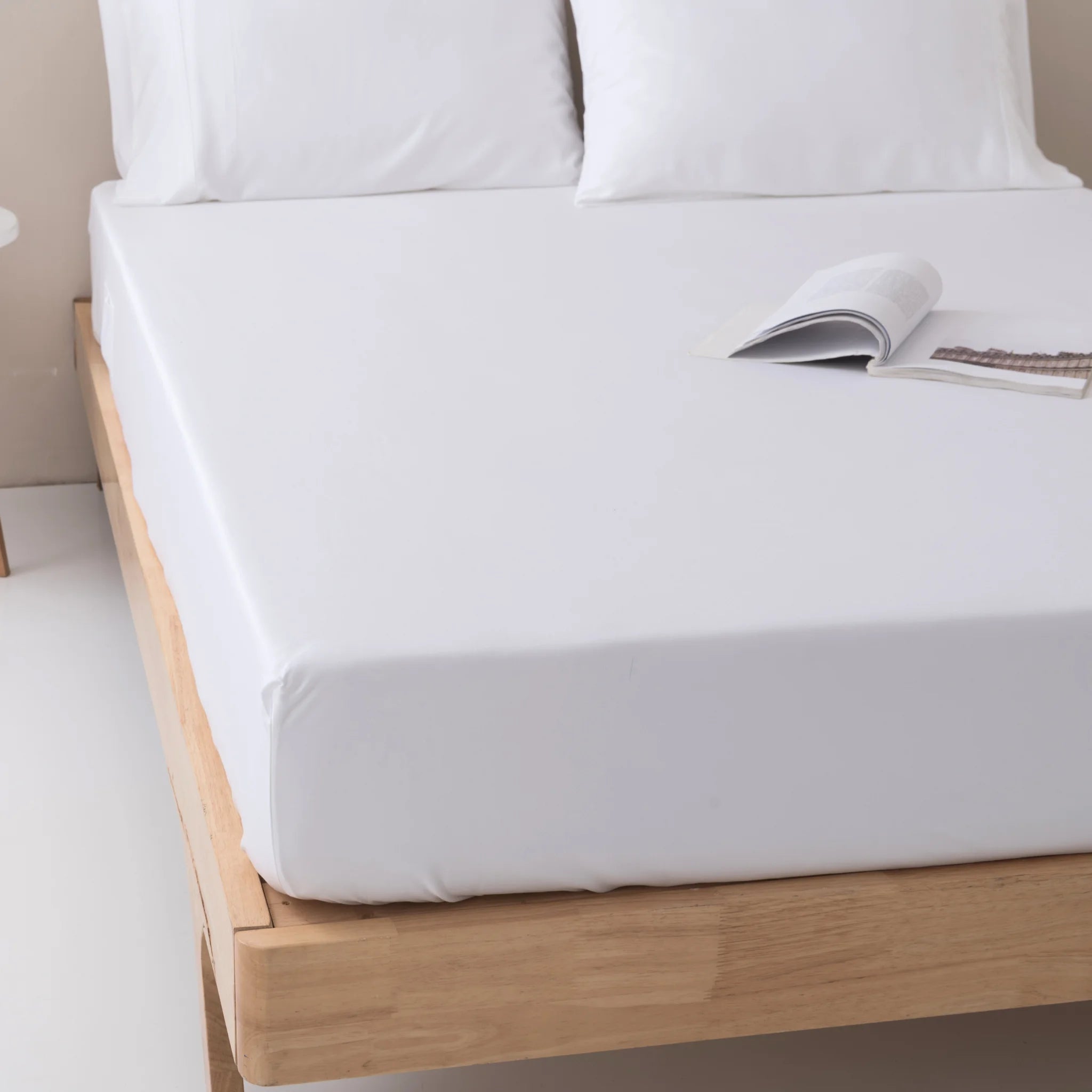 A neatly made wooden bed features the Linenly Bamboo Fitted Sheet in white with deep sides and plush pillows, while an open book rests near the pillows.