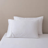 Two white Linenly Bamboo Pillowcase Set pillows rest on a neatly made bed with a fitted white sheet, set against a plain, light beige wall.