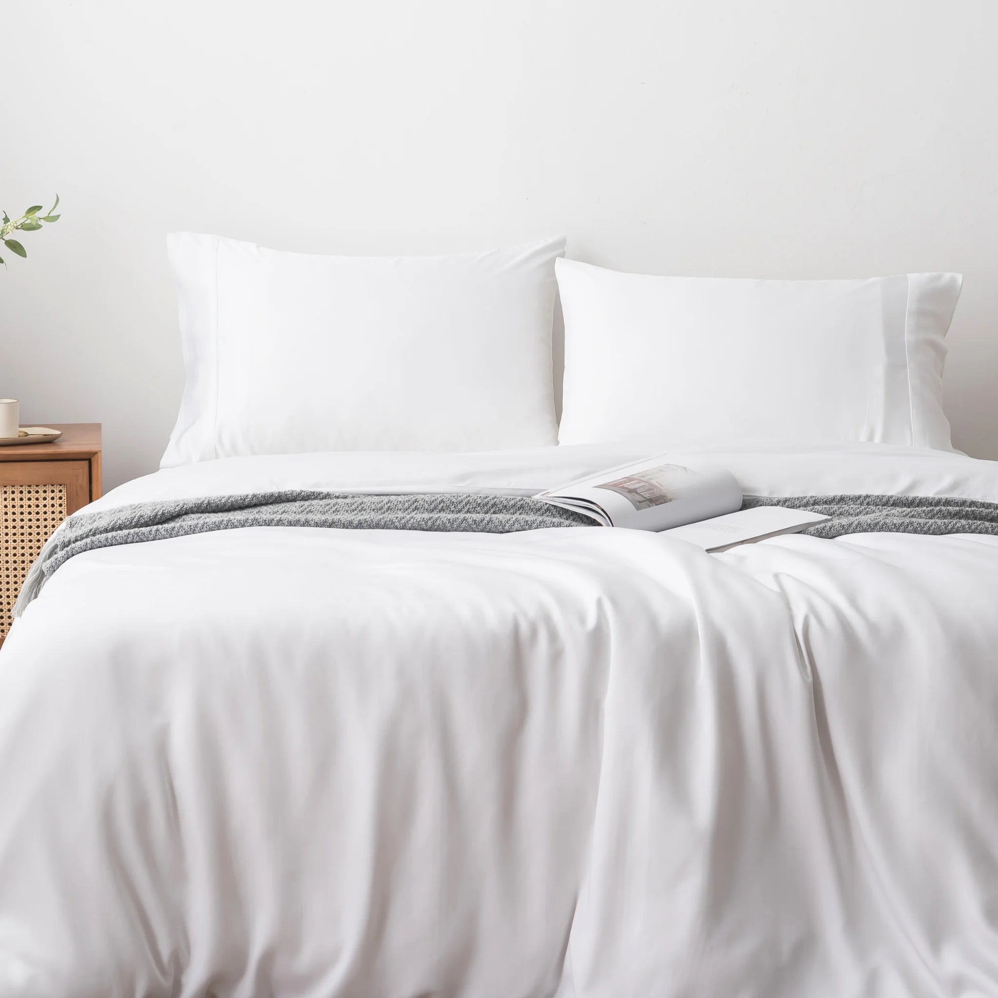 A neatly made bed with white sheets, two pillows, a gray patterned blanket at the foot, and an open book on the duvet features the Linenly Bamboo Quilt Cover Set - White for a luxurious touch.