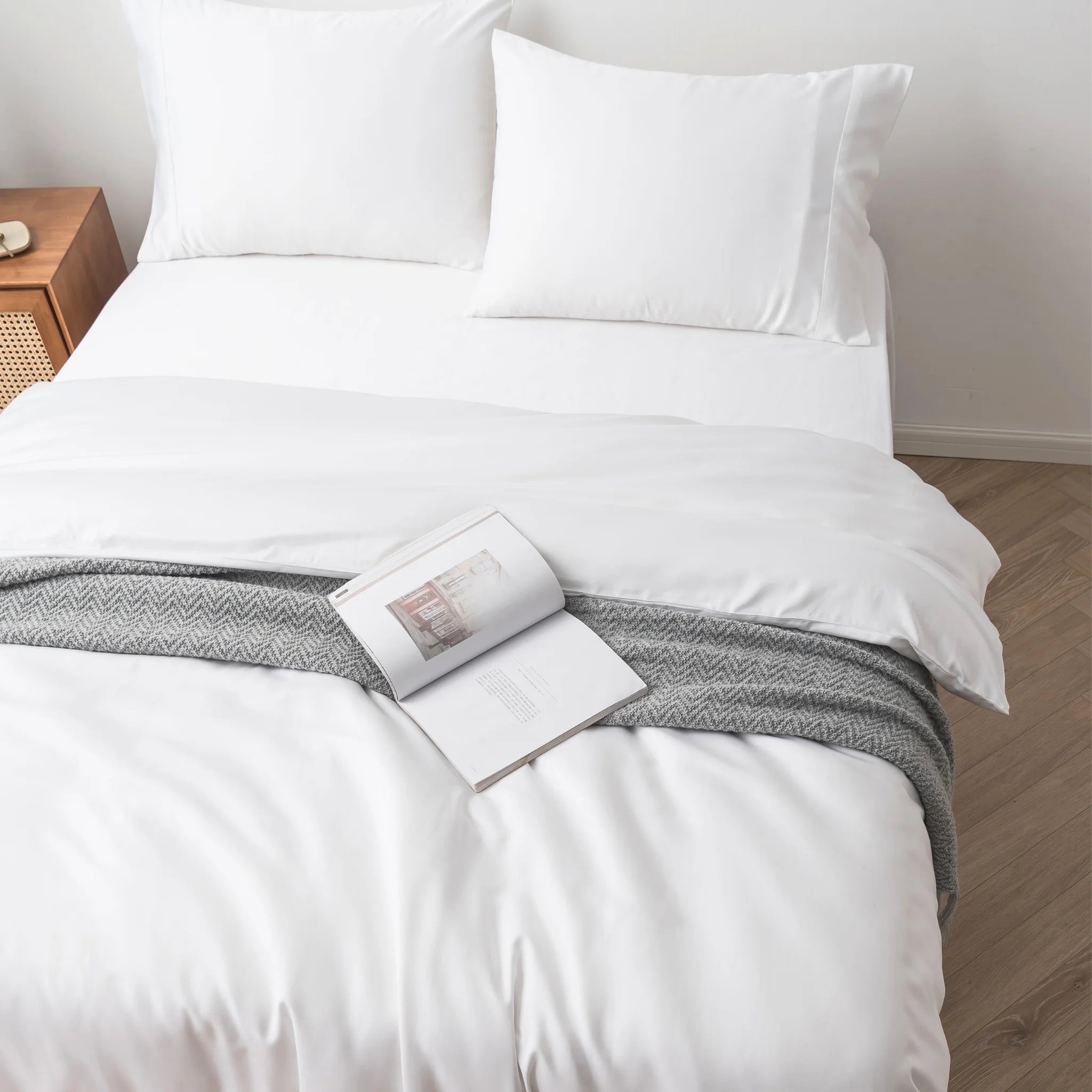 A neatly made bed features the Linenly Bamboo Quilt Cover Set - White in a soft sateen weave, with white sheets and pillows, a gray patterned throw, and an open book on top. A wooden nightstand sits beside the bed on a light-colored floor.