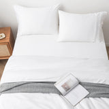 A neatly made bed with white sheets and two pillows sits by a wooden nightstand. A gray patterned blanket, similar to the Linenly Bamboo Quilt Cover Set - White, is folded at the foot with an open magazine on top.