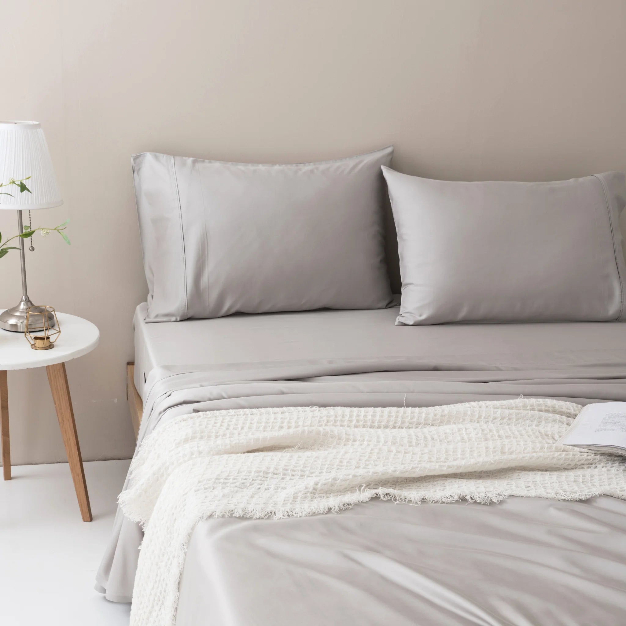 A bed is made with Linenly’s Bamboo Sheet Set in Silver and two pillows, topped with a textured cream blanket. A white nightstand holds a lamp, small plant, and water glass. An open book rests on the luxury sheets.