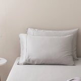 Two neatly arranged white pillows rest against a light wall on a made bed dressed in Linenly’s Bamboo Sheet Set - Silver; a small round bedside table is partially visible on the left.