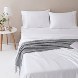 A neatly made bed features Linenly's ultra-soft Bamboo Sheet Set in White, two white pillows, and a gray knitted blanket. Next to the bed is a round white nightstand with a lamp, green branches in a glass vase, and a small candle.