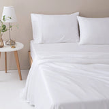A neatly made bed features the Linenly Bamboo Sheet Set - White and two white pillows. A round, white side table holds a lamp, small plant in a glass vase, and a water glass, completing the minimalist room style.