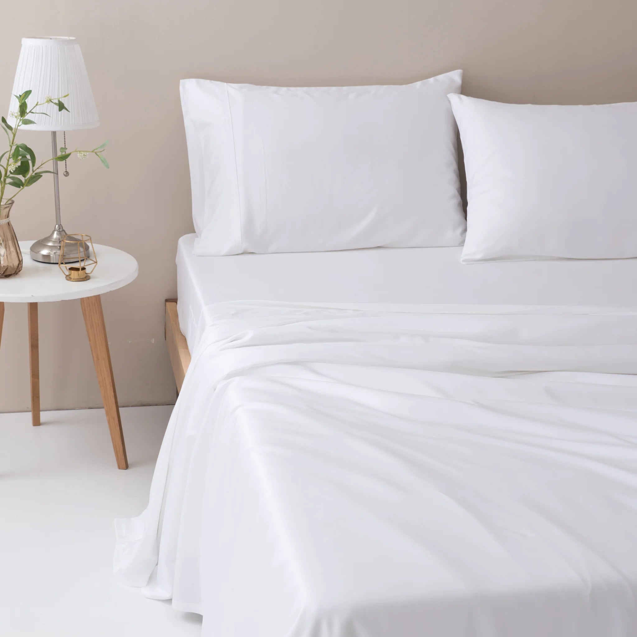 A neatly made bed features the Linenly Bamboo Sheet Set - White and two white pillows. A round, white side table holds a lamp, small plant in a glass vase, and a water glass, completing the minimalist room style.